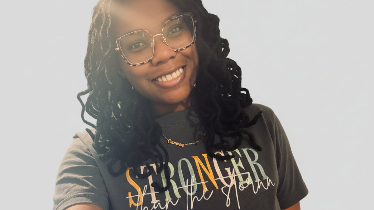 A smiling woman with long black curly hair and square tortoiseshell glasses takes a selfie while wearing a dark gray graphic t-shirt that reads 'STRONGER than the storm' in yellow, orange, and white lettering.